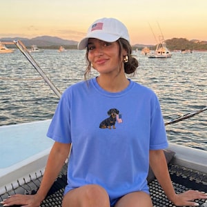 Op de afbeelding: Een lichtblauw T-shirt met een geborduurde teckel met een kleine Amerikaanse vlag. De persoon draagt een witte baseballcap met een Amerikaanse vlag. De achtergrond toont water en boten.