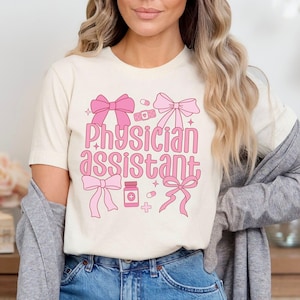May include: A white t-shirt with a pink graphic design that says "Physician Assistant" with pink bows, pills, and bandages.