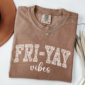 Op de afbeelding: Een bruin Comfort Colors T-shirt met de woorden "FRI-YAY vibes" in witte bloemenletters. Een zilveren ketting met een kleine hanger ligt over het shirt. Een bruine hoed is zichtbaar in de linkerbovenhoek.
