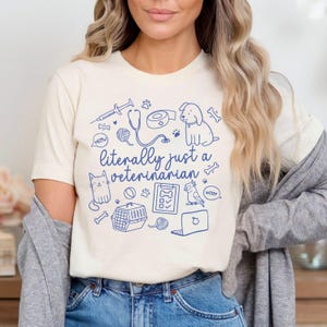 May include: A white t-shirt with a blue graphic design that says "literally just a veterinarian". The graphic includes a stethoscope, a syringe, a cat, a dog, a bird, a bone, a ball, a pet carrier, a leash, a bandage, and a laptop.