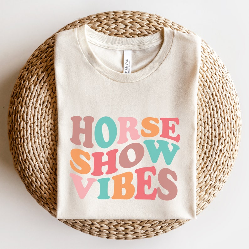May include: A beige t-shirt with the text "HORSE SHOW VIBES" in colorful, retro-style lettering.
