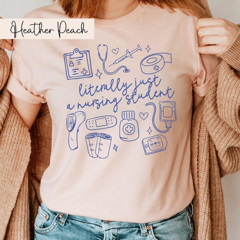 May include: A pink t-shirt with the text "literally just a nursing student" in blue. The shirt features a variety of medical illustrations, including a stethoscope, a thermometer, a bandage, a pill bottle, and a blood glucose monitor.