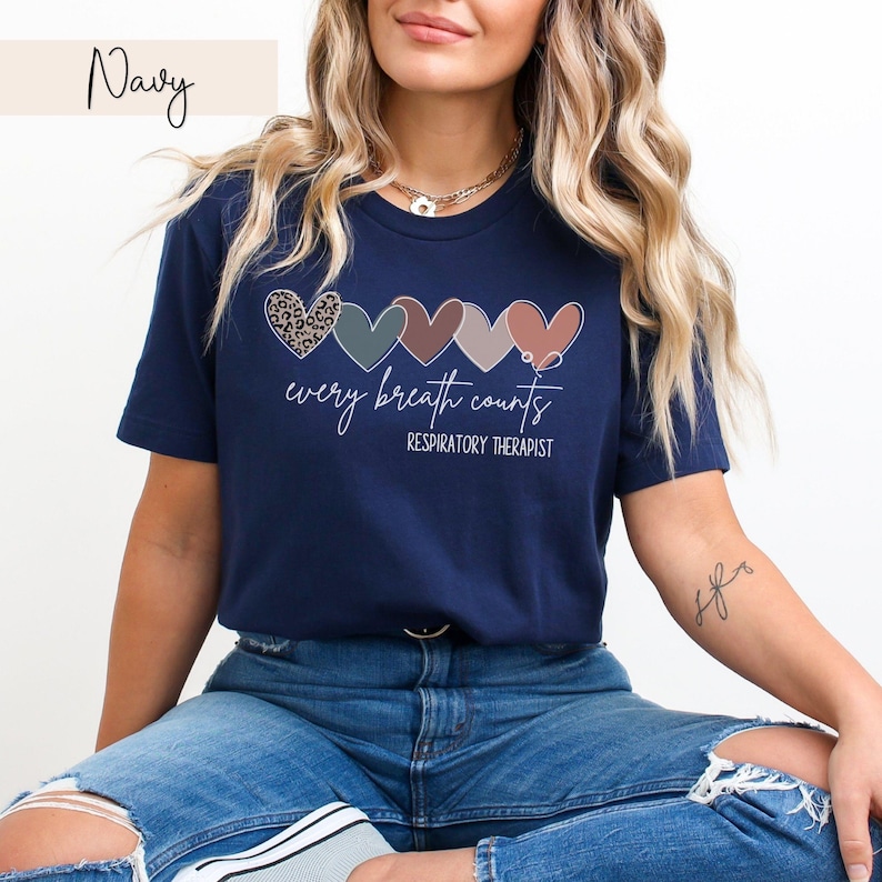 以下が含まれることがあります： 5つの異なる模様のカラフルなハートが並んだネイビーブルーのTシャツ。ハートの下には「every breath counts」というテキストが印刷され、その下に「RESPIRATORY THERAPIST」という単語が印刷されています。