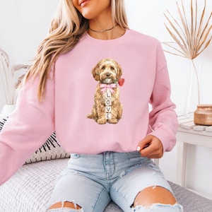 May include: A pale pink sweatshirt with a watercolour illustration of a dog holding a red rose. The dog has a pink bow and the word "LOVE" is printed vertically in front of it. The sweatshirt is a plain colour.