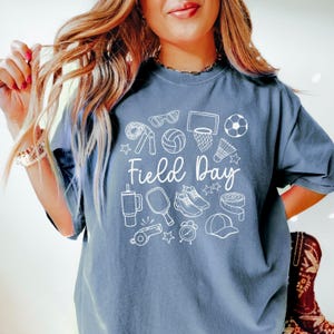 May include: A blue t-shirt with a white graphic design of various sports equipment and the text "Field Day".