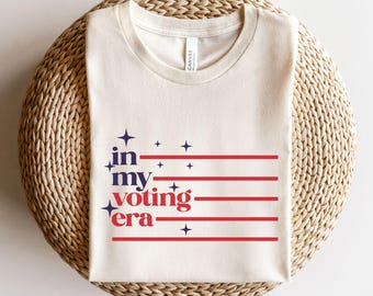 May include: A beige t-shirt with the text "in my voting era" printed in blue and red. The text is surrounded by small stars and red and white stripes.