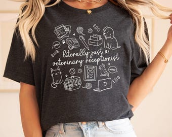 Könnte beinhalten: Ein dunkelgraues T-Shirt mit einem weißen Grafikdesign, das das Arbeitsleben einer Tierarzt-Rezeptionistin darstellt. Das Design beinhaltet eine Katze, einen Hund, einen Vogel, einen Computer und andere Gegenstände, die mit der tierärztlichen Versorgung zusammenhängen. Der Text auf dem Shirt lautet "literally just a veterinary receptionist".