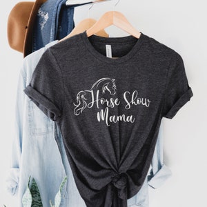 Peut inclure: T-shirt gris fonc&eacute; avec des manches retrouss&eacute;es et le texte "Horse Show Mama" en &eacute;criture blanche. Un dessin de cheval blanc est au-dessus du texte. Le t-shirt est suspendu &agrave; un cintre en bois avec d'autres v&ecirc;tements.