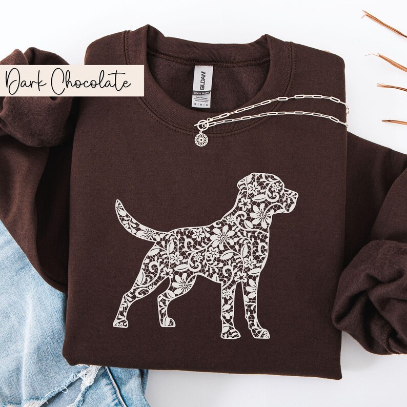 May include: Dark chocolate-colored sweatshirt featuring a white floral lace design of a dog. The sweatshirt has a crew neck and long sleeves. A silver necklace with a pendant is draped on the sweatshirt. The words "Dark Chocolate" are in a white banner.