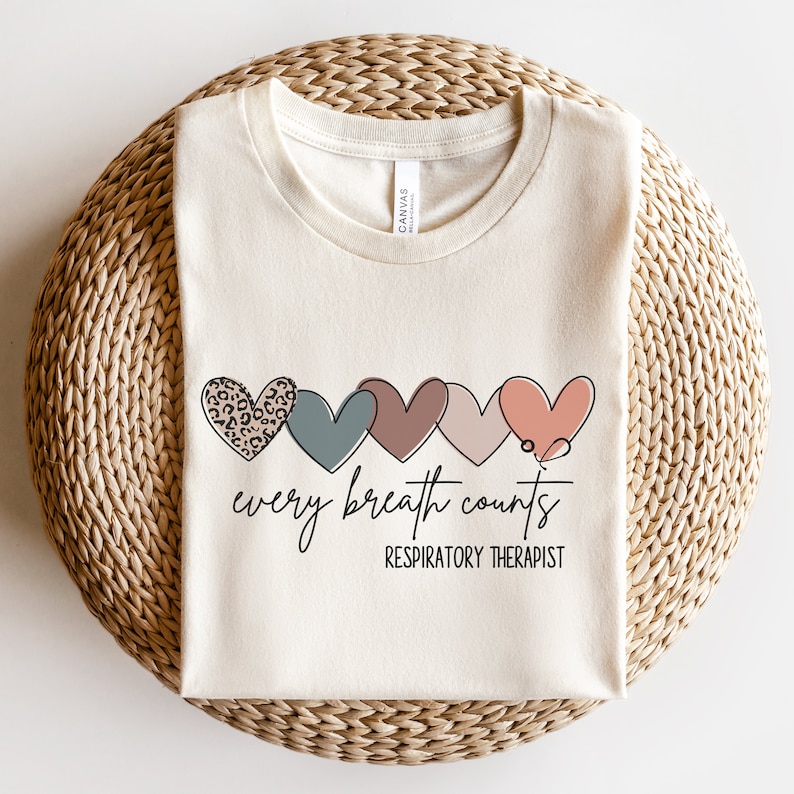 以下が含まれることがあります： ベージュのTシャツに、5つのハートが横に並んだデザインが施されています。ハートはそれぞれ異なる色と模様で、レオパード柄も含まれています。ハートの下には「every breath counts」というテキストが、その下には「RESPIRATORY THERAPIST」というテキストが書かれています。