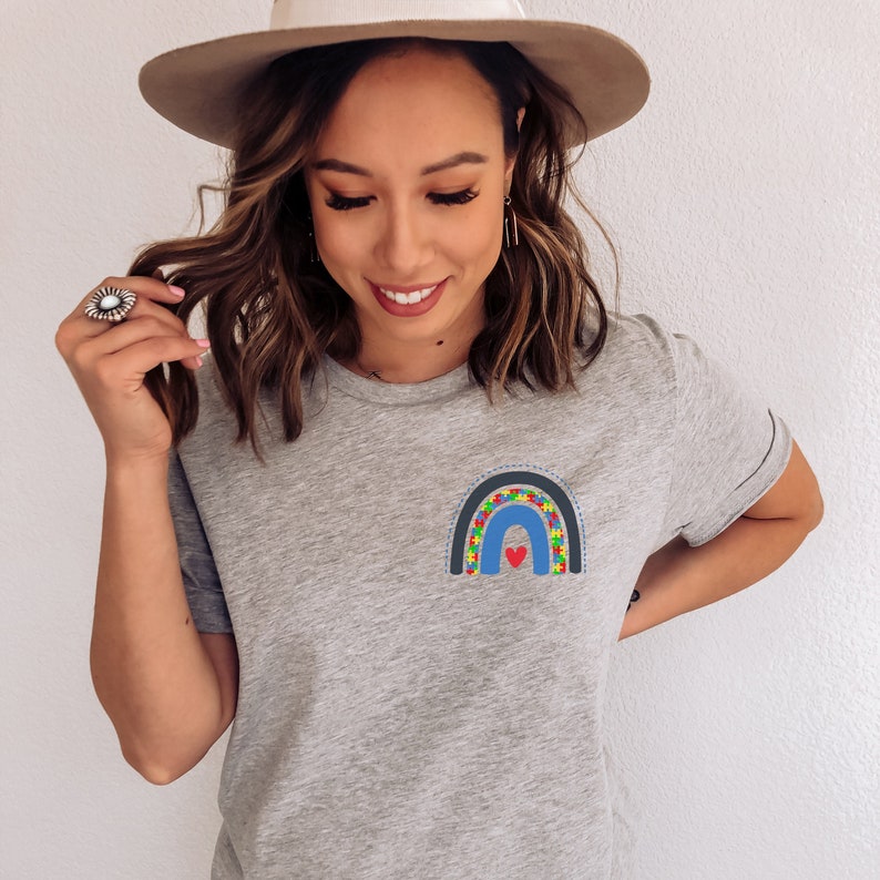 Op de afbeelding: Een grijs T-shirt met een regenboogontwerp gemaakt van puzzelstukjes. De regenboog heeft een rood hart in het midden.