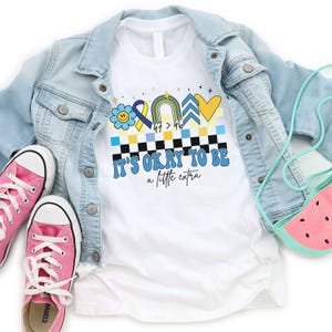 May include: White t-shirt with a blue, yellow, and rainbow design. The design includes a smiley face, a ribbon, a rainbow, an arrow, and a heart. The text "It's okay to be a little extra" is printed below the design.