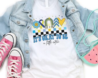 Può includere: Maglietta bianca con un design blu, giallo e arcobaleno. Il design include una faccia sorridente, un nastro, un arcobaleno, una freccia e un cuore. Il testo "It's okay to be a little extra" è stampato sotto il design.