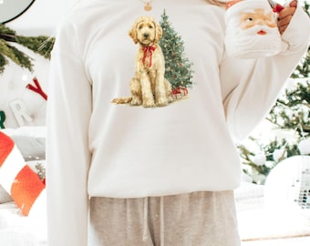 May include: White sweatshirt featuring a watercolor illustration of a golden doodle dog with a red bow, sitting next to a Christmas tree. The dog is surrounded by small wrapped gifts. A Santa mug is held in the right hand.