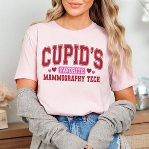 May include: A pale pink t-shirt featuring the maroon text "CUPID'S FAVORITE MAMMOGRAPHY TECH". The text is in a collegiate style font with heart symbols.