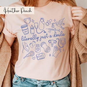 May include: Heather Peach t-shirt featuring blue line art illustrations of baby-related items and the text "literally just a doula". The design includes a stork, baby bottle, and medical equipment.