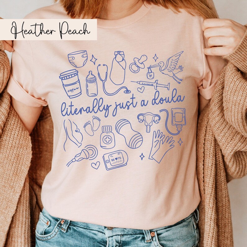 May include: Heather Peach t-shirt featuring blue line art illustrations of baby-related items and the text "literally just a doula". The design includes a stork, baby bottle, and medical equipment.
