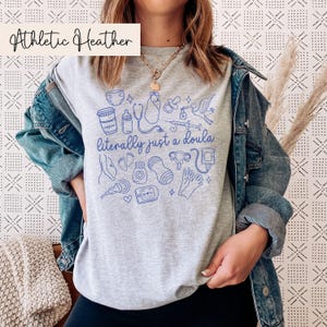 May include: A grey Athletic Heather t-shirt featuring a blue illustration of baby-related items and the text "literally just a doula." The shirt has a crew neck and short sleeves. The text "Athletic Heather" is in a white box in the upper left corner.