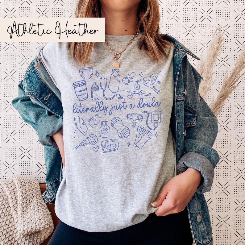 May include: A grey Athletic Heather t-shirt featuring a blue illustration of baby-related items and the text "literally just a doula." The shirt has a crew neck and short sleeves. The text "Athletic Heather" is in a white box in the upper left corner.