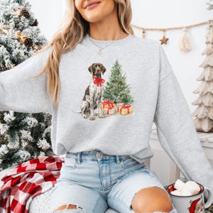 May include: A light gray sweatshirt featuring a watercolor illustration of a German Shorthaired Pointer wearing a red bow, a Christmas tree, and wrapped gifts. The dog is brown and white. A festive holiday design.
