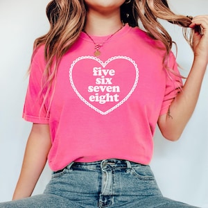 May include: A pink t-shirt with a white heart graphic that says "five six seven eight" in a retro font.