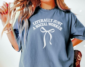 Op de afbeelding: Een blauw T-shirt met de tekst "Literally Just A Social Worker" in wit met een witte strik eronder.