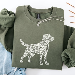 May include: An olive green sweatshirt with a white floral dog silhouette design. A silver necklace with a small pendant rests on the sweatshirt. The sweatshirt is folded, with the sleeves and collar visible.