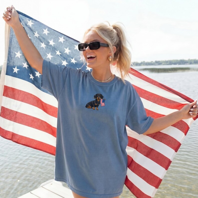 Op de afbeelding: Een blauw t-shirt met een geborduurde teckel met een kleine Amerikaanse vlag. De persoon houdt een grote Amerikaanse vlag vast. Het shirt heeft een effen blauwe kleur.