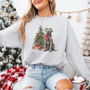 May include: A light gray sweatshirt featuring a watercolor illustration of a dog with a red bow, a Christmas tree, and wrapped gifts. The dog is sitting in front of the tree. The sweatshirt is a casual, festive garment.
