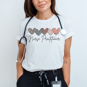 Op de afbeelding: Wit T-shirt met een ontwerp van vijf harten in verschillende kleuren en patronen, waaronder een hart met luipaardprint. De woorden "Nurse Practitioner" staan in een scriptletter onder de harten. Een stethoscoop hangt om de nek.