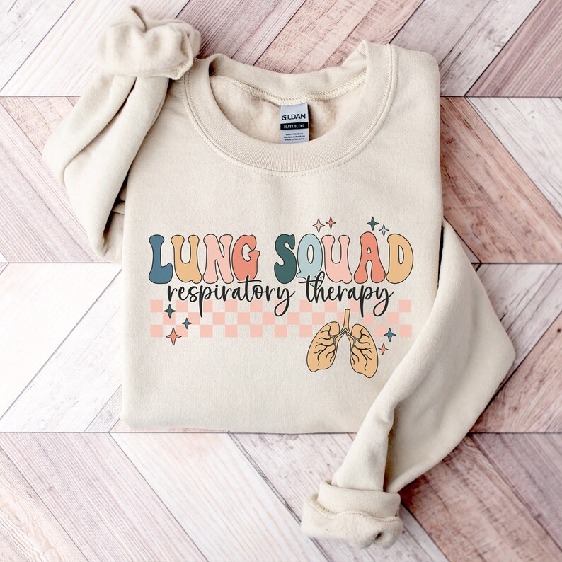 May include: A beige sweatshirt with the text "LUNG SQUAD respiratory therapy" in colorful letters. The text is surrounded by a checkered pattern and stars. A cartoon image of lungs is below the text.