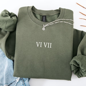 May include: An olive green sweatshirt with the Roman numerals VI VII printed in white. A silver necklace with a circular pendant is draped over the sweatshirt. The sweatshirt is folded, with the sleeves and a portion of blue jeans visible.