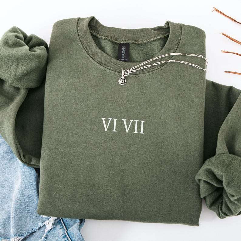 May include: An olive green sweatshirt with the Roman numerals VI VII printed in white. A silver necklace with a circular pendant is draped over the sweatshirt. The sweatshirt is folded, with the sleeves and a portion of blue jeans visible.