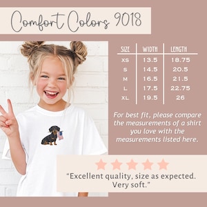 Op de afbeelding: Wit Comfort Colors T-shirt met een teckel met een Amerikaanse vlag. De maattabel toont de afmetingen in centimeters. De tekst "Comfort Colors 9018" staat bovenaan. Het shirt wordt beschreven als "Uitstekende kwaliteit, maat zoals verwacht. Zeer zacht."