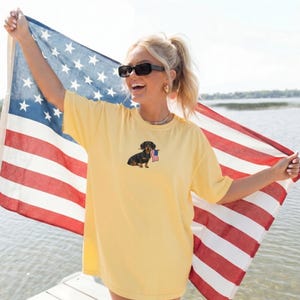 Op de afbeelding: Lichtgeel T-shirt met een teckel die een kleine Amerikaanse vlag vasthoudt. De persoon houdt een Amerikaanse vlag op de achtergrond vast. Het shirt heeft een lichtgele kleur.