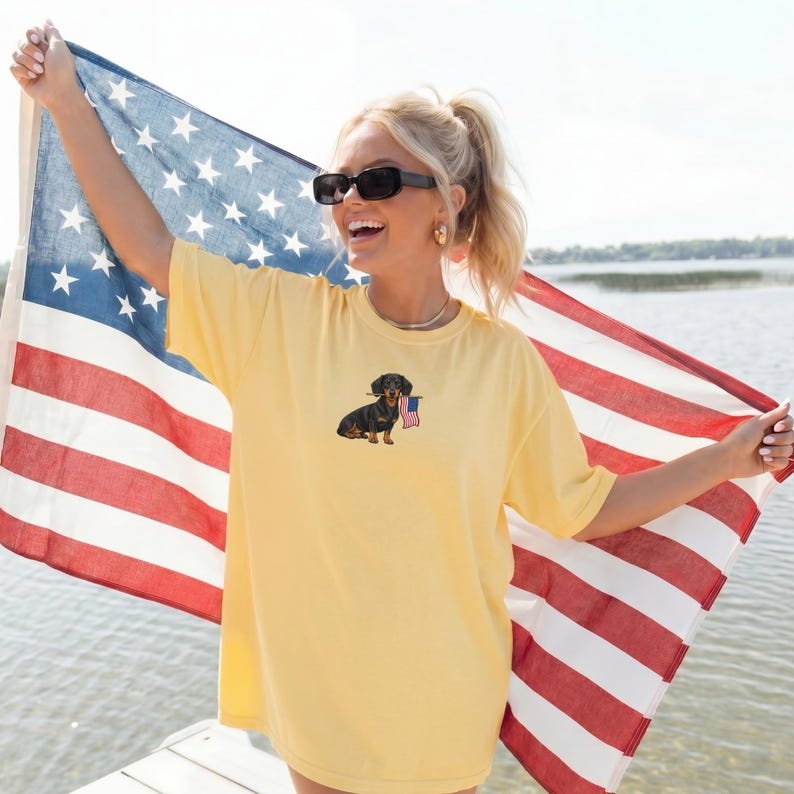 Op de afbeelding: Lichtgeel T-shirt met een teckel die een kleine Amerikaanse vlag vasthoudt. De persoon houdt een Amerikaanse vlag op de achtergrond vast. Het shirt heeft een lichtgele kleur.