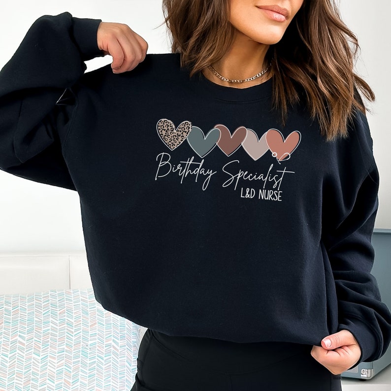 Op de afbeelding: Zwart sweatshirt met een afbeelding van vijf kleurrijke harten en de tekst "Birthday Specialist L&D NURSE"