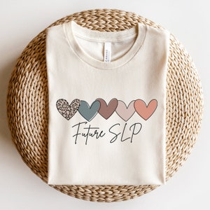May include: A beige t-shirt with a line of five hearts in different colors and patterns. The hearts are a leopard print, teal, brown, pink, and coral. The text "Future SLP" is written in black cursive below the hearts.