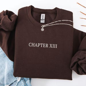 May include: A dark brown sweatshirt with the text "CHAPTER XIII" in white. A silver necklace with a circular pendant is draped across the sweatshirt. The sweatshirt is folded, with a portion of blue jeans visible.