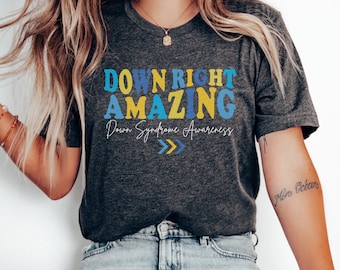 May include: A dark gray t-shirt with the text "Down Right Amazing" in blue, yellow, and white letters. The text "Down Syndrome Awareness" is written in cursive below the main text. Two yellow arrows point to the right.