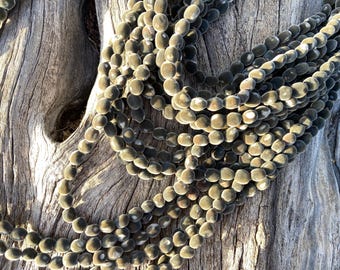 May include: Strands of dark, oval-shaped beads with a velvety texture are draped over a weathered, light-brown wooden surface. The beads are a muted olive-green color, and the background is a natural wood grain.