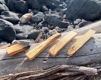 Puede incluir: Cuatro palitos de madera, algunos con un acabado ligeramente quemado, están dispuestos sobre un tronco gris desgastado. Los palitos son de color marrón claro y tienen una veta de madera natural. El tronco está apoyado sobre un lecho de rocas volcánicas negras.