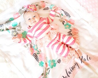 May include: A white baby blanket with a pink and green floral design. The blanket has a pink ribbon with the name "Delaney Kate" written in black cursive.