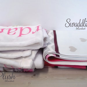 May include: Two plush baby blankets stacked on top of each other. The top blanket is white with pink lettering. The second blanket is white with a pattern of hearts and a chevron design. The words "Swaddling blanket" and "Plush blanket" are visible.