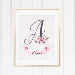 May include: A framed print with a white woodgrain background featuring a large cursive letter A with pink and purple flowers and feathers. A pink ribbon banner with the name "Alana" is below the letter.