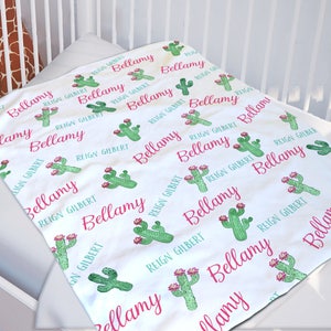 May include: White baby blanket with a repeating pattern of green cactus plants and the names "Bellamy" and "Reign Gilbert" in pink lettering.