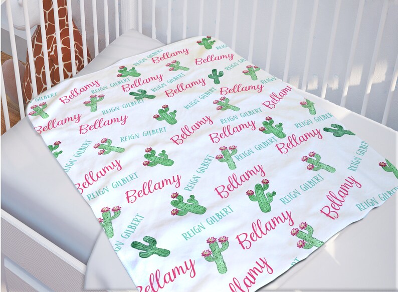 May include: White baby blanket with a repeating pattern of green cactus plants and the names "Bellamy" and "Reign Gilbert" in pink lettering.
