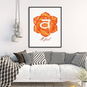 May include: A framed print featuring an orange watercolor flower with a white center and the word "I feel" in script. The artwork hangs above a white sofa with patterned pillows and a textured throw blanket.