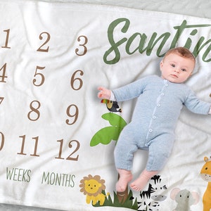 K&ouml;nnte beinhalten: Wei&szlig;e Meilenstein-Decke mit braunen Zahlen 1-12, den W&ouml;rtern "TAGE", "WOCHEN" und "MONATE" sowie dem Namen "Santino" in Gr&uuml;n. Die Decke zeigt Dschungeltier-Illustrationen und ein Baby in einem blauen Strampler.