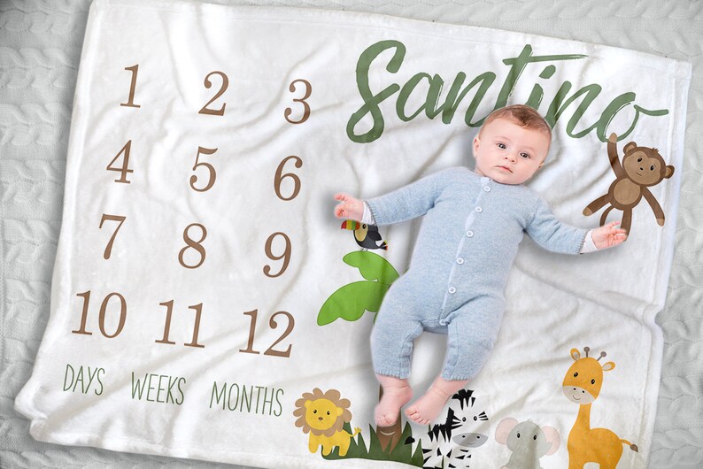 K&ouml;nnte beinhalten: Wei&szlig;e Meilenstein-Decke mit braunen Zahlen 1-12, den W&ouml;rtern "TAGE", "WOCHEN" und "MONATE" sowie dem Namen "Santino" in Gr&uuml;n. Die Decke zeigt Dschungeltier-Illustrationen und ein Baby in einem blauen Strampler.